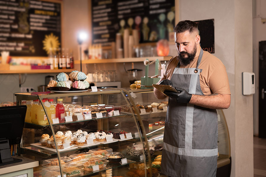 Successful small business owner in cafe using digital tablet and looking at screen. Serious barista with a beard standing in front of showcase while counting, Start up coffee shop