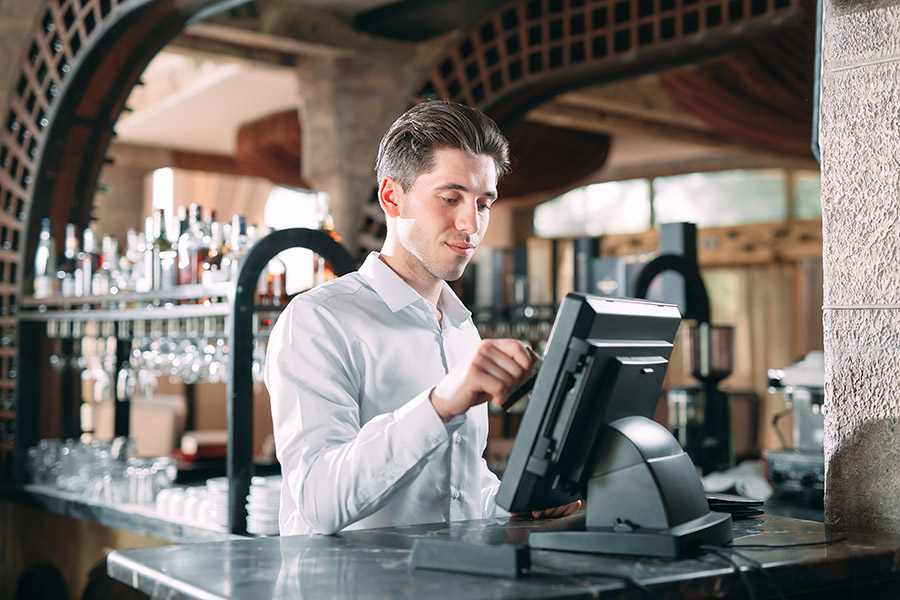 small business, people and service concept - happy man or waiter in apron at counter with cashbox working at bar or coffee shop